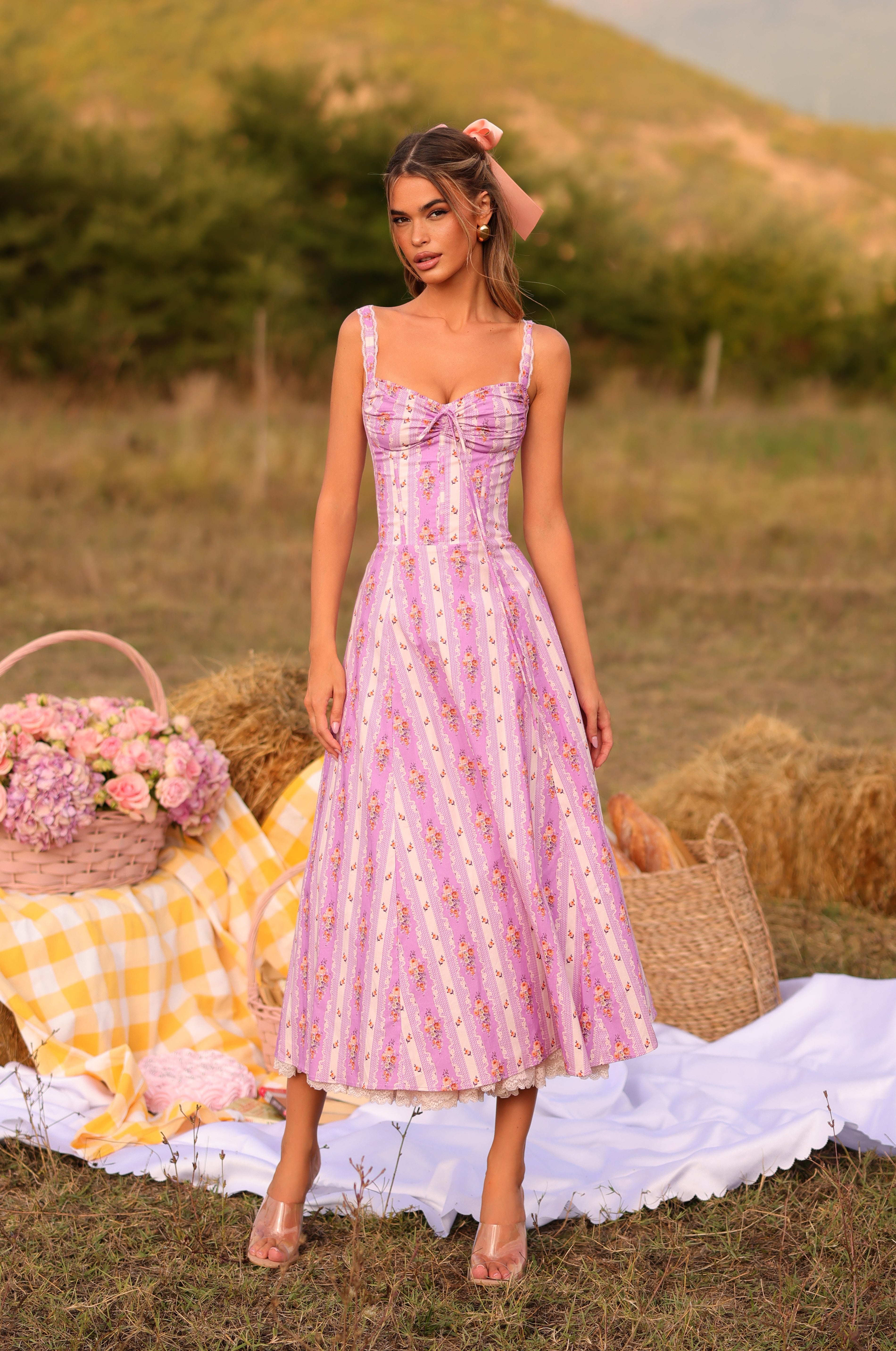 A woman wears the Lilac Floral Casual Midi Dress: Celenia, standing on a picnic setup with a basket, flowers, and checkered blanket—an ideal summer wedding guest dress for a grassy outdoor event.